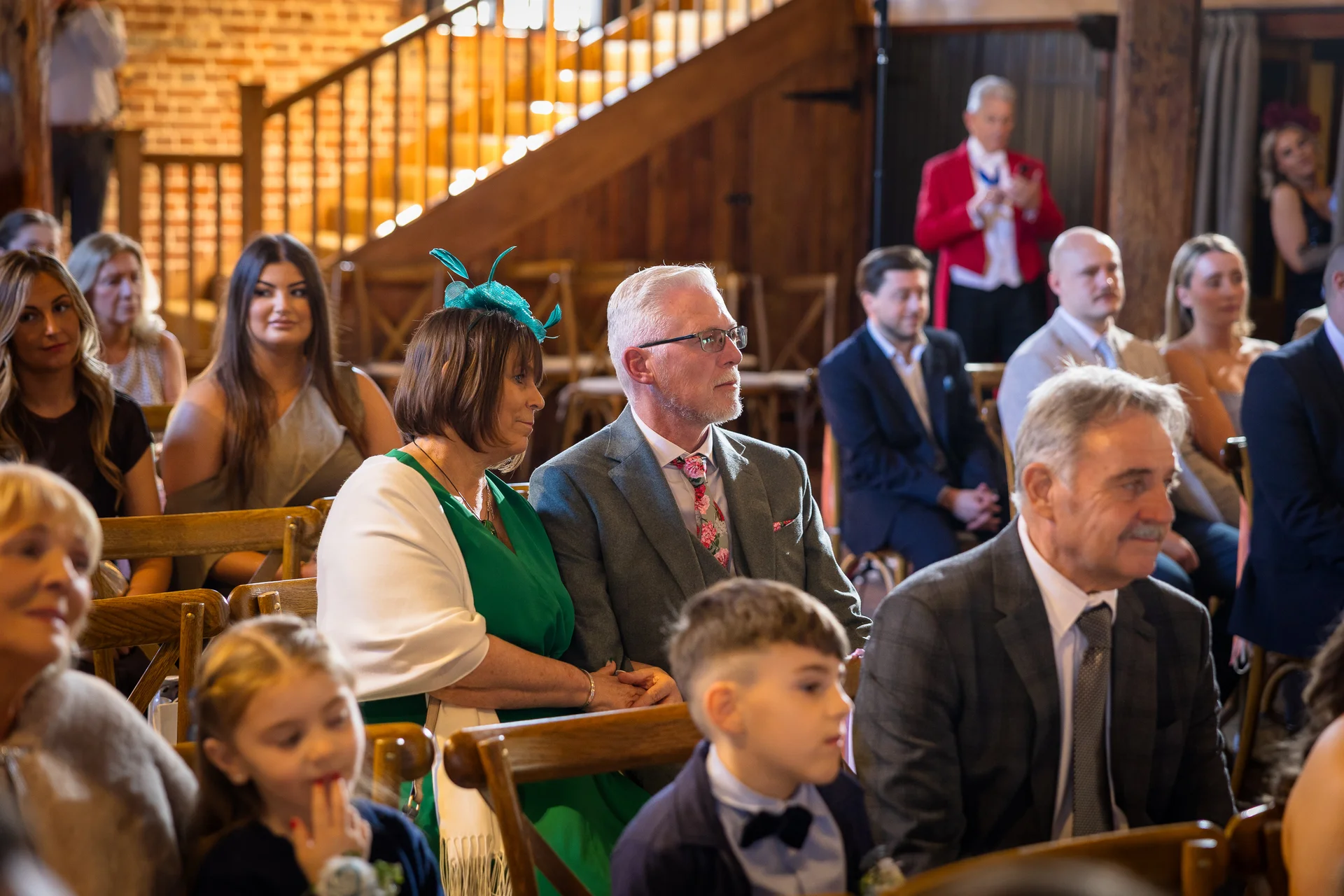 Guests seated on wooden chairs inside a rustic barn wedding venue, watching the ceremony unfold under exposed wooden rafters.