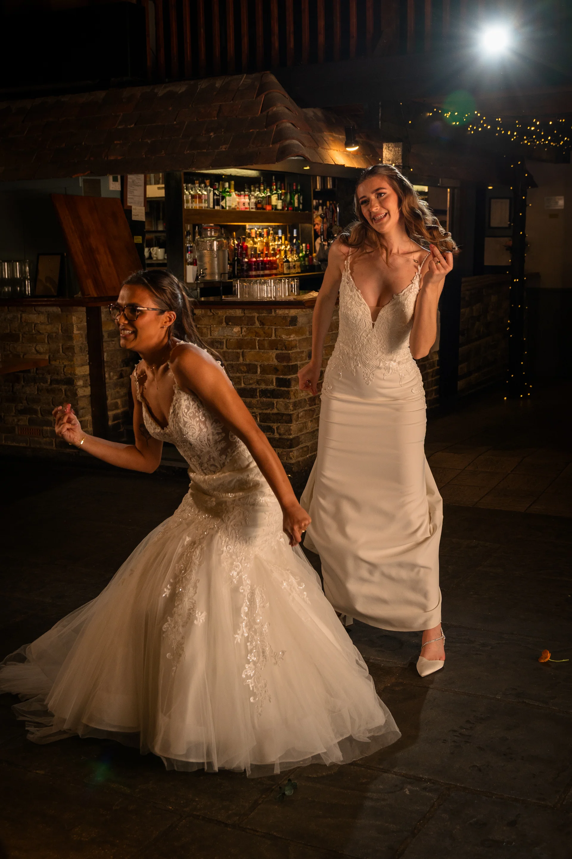 Two smiling brides in gorgeous white wedding dresses joyfully dance together inside a rustic pub venue during their evening reception.