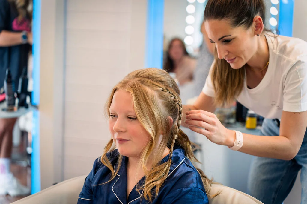 A young bridesmaid sits calmly while a hairstylist expertly braids her blonde hair during morning wedding preparations.