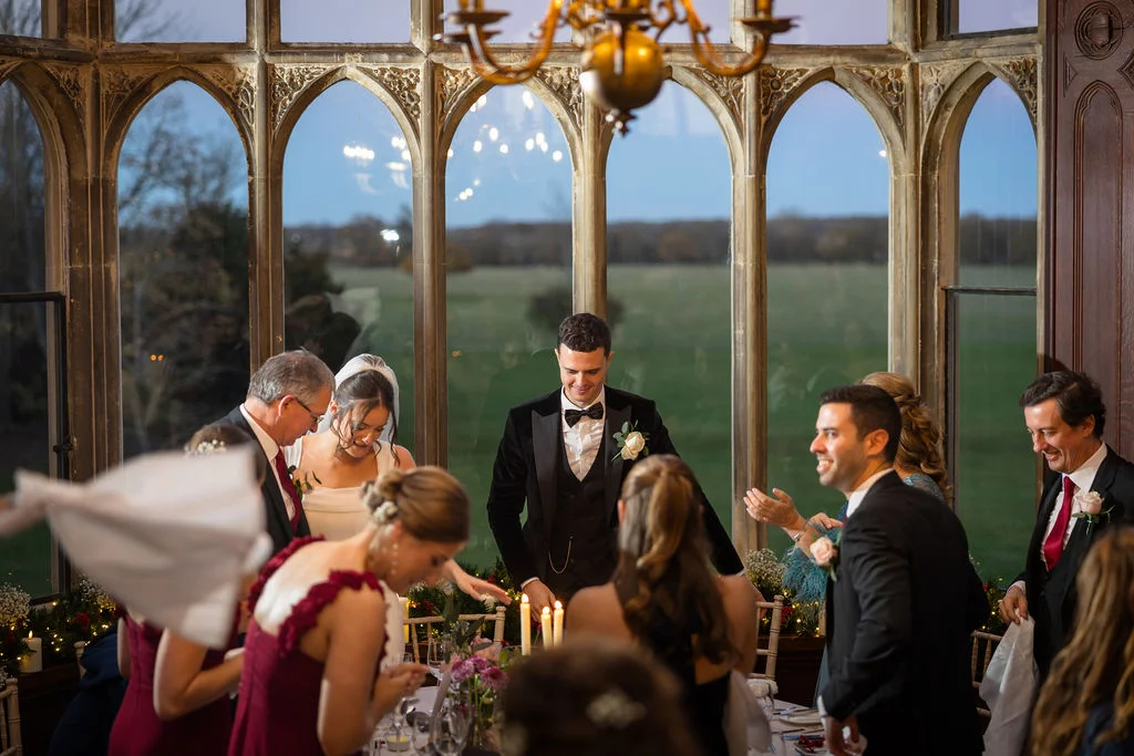 The bride and groom stand smiling at the top table in a grand room with large arched windows during evening wedding speeches.