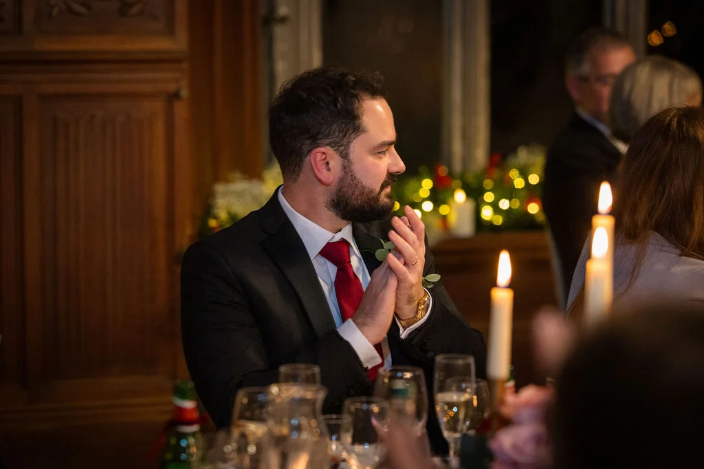 A groom in a black suit and red tie sits at a candlelit reception table, clapping and laughing during the wedding speeches.