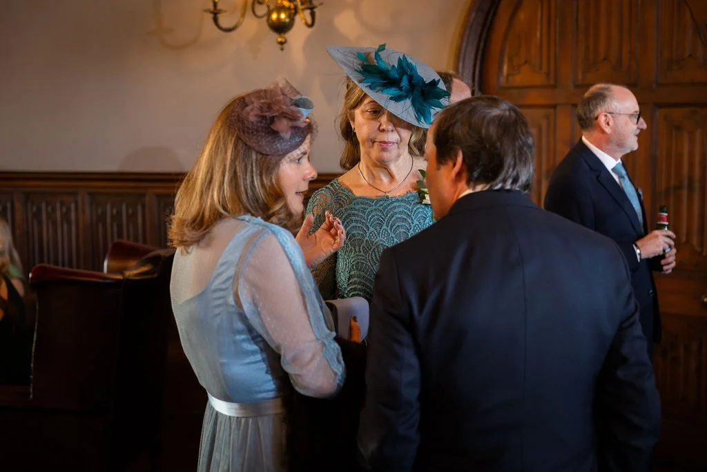 Wedding guests wearing elegant hats and formal attire mingle and chat in a wood-panelled reception room.