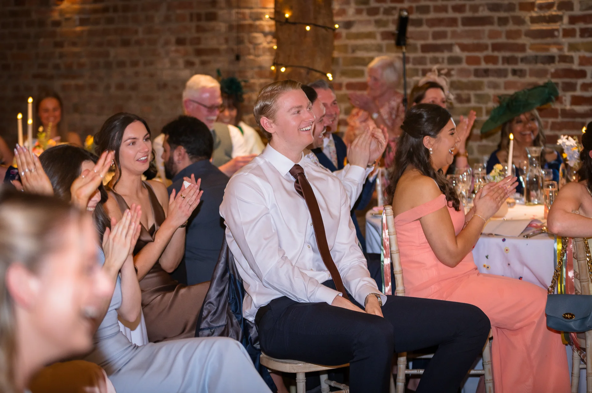 Wedding guests sit at tables in a rustic brick barn venue, smiling and applauding during the reception speeches.