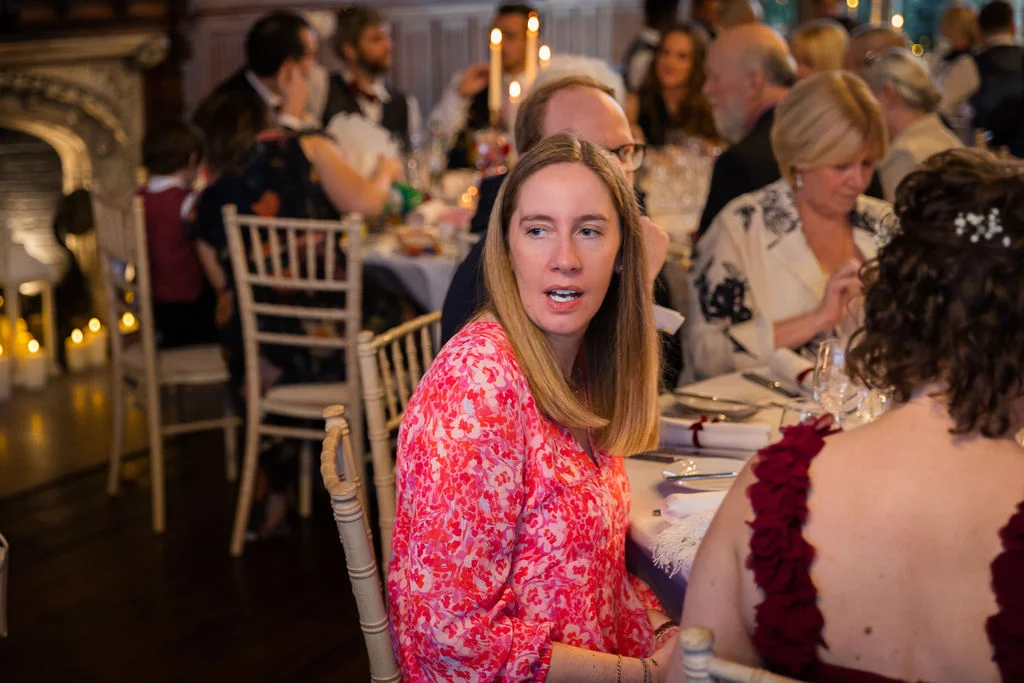 A guest wearing a bright pink floral print dress is engaged in conversation at a candlelit wedding reception table.