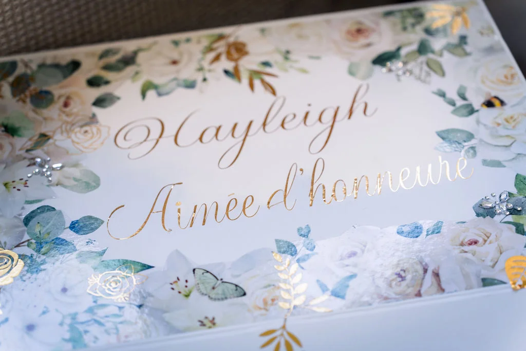 A close-up shot of a personalised wedding box decorated with soft floral illustrations, greenery, and elegant gold calligraphy.