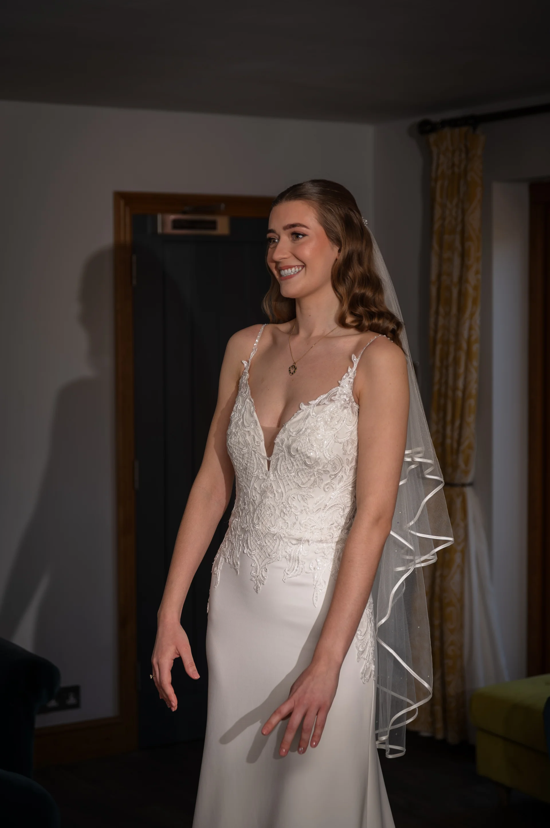 A smiling bride wearing a v-neckline crepe wedding dress with lace details and a ribbon-trimmed veil stands indoors during bridal preparation.