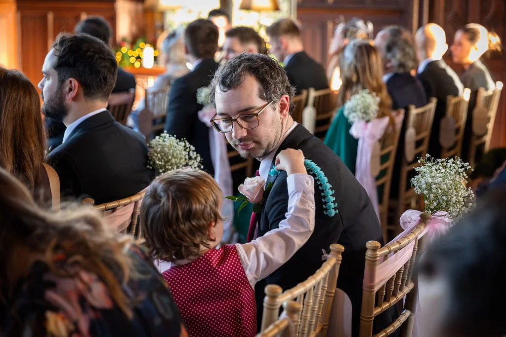 A young page boy in a red polka-dot waistcoat touches a guest's shoulder inside a wood-panelled wedding ceremony room.