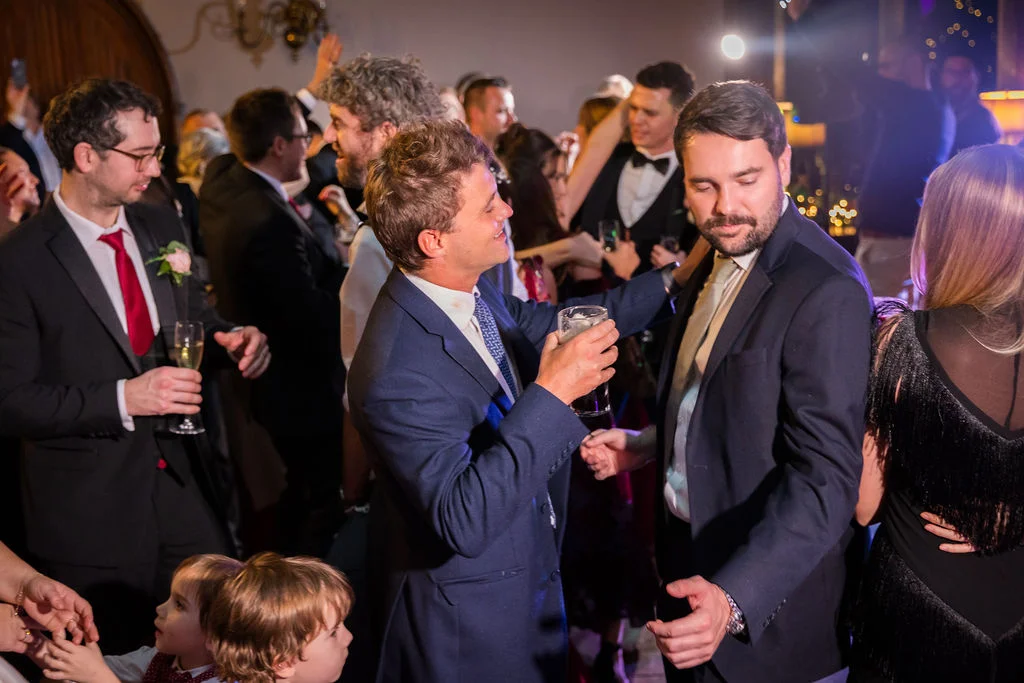 A lively wedding reception where smartly dressed guests in suits mingle and celebrate on a crowded dance floor.