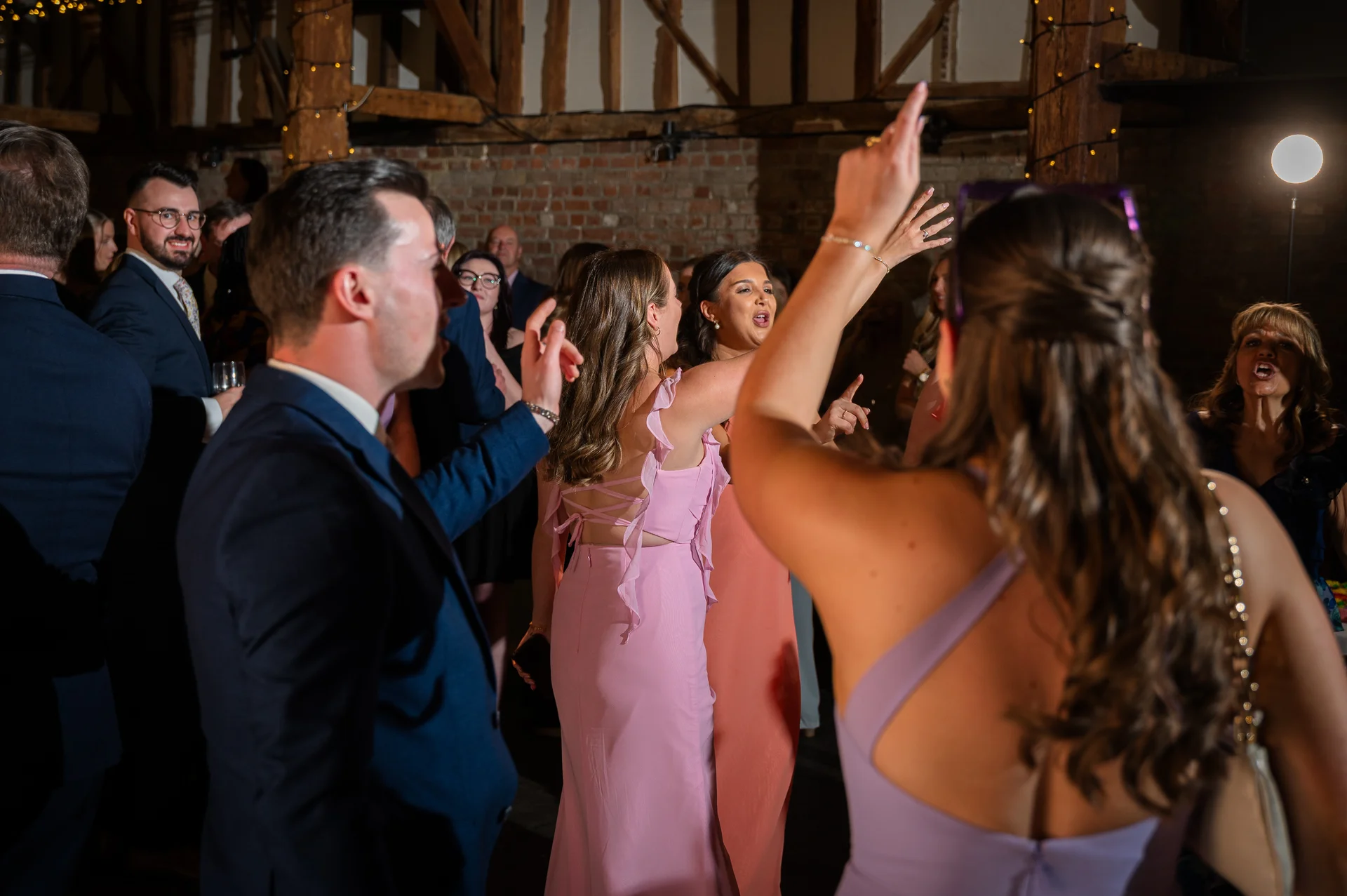 Wedding guests enjoy the evening reception on the dance floor inside a rustic converted barn with exposed oak beams and brickwork.