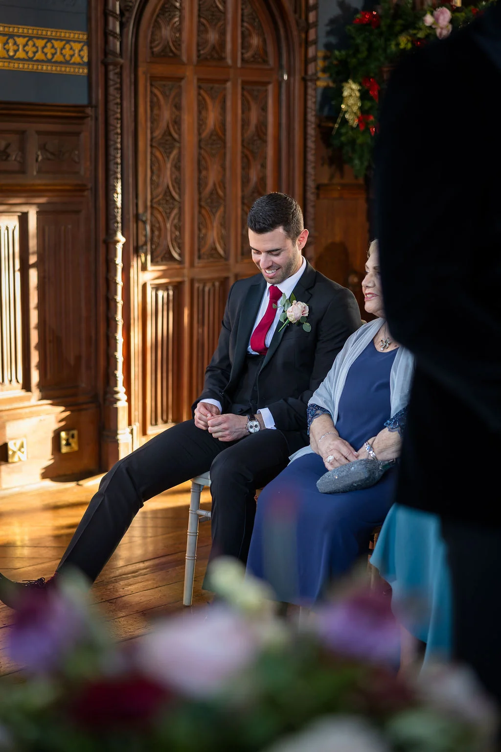 A groom in a black three-piece suit with a red tie and buttonhole waits inside a wood-panelled room, seated next to an elderly family member.