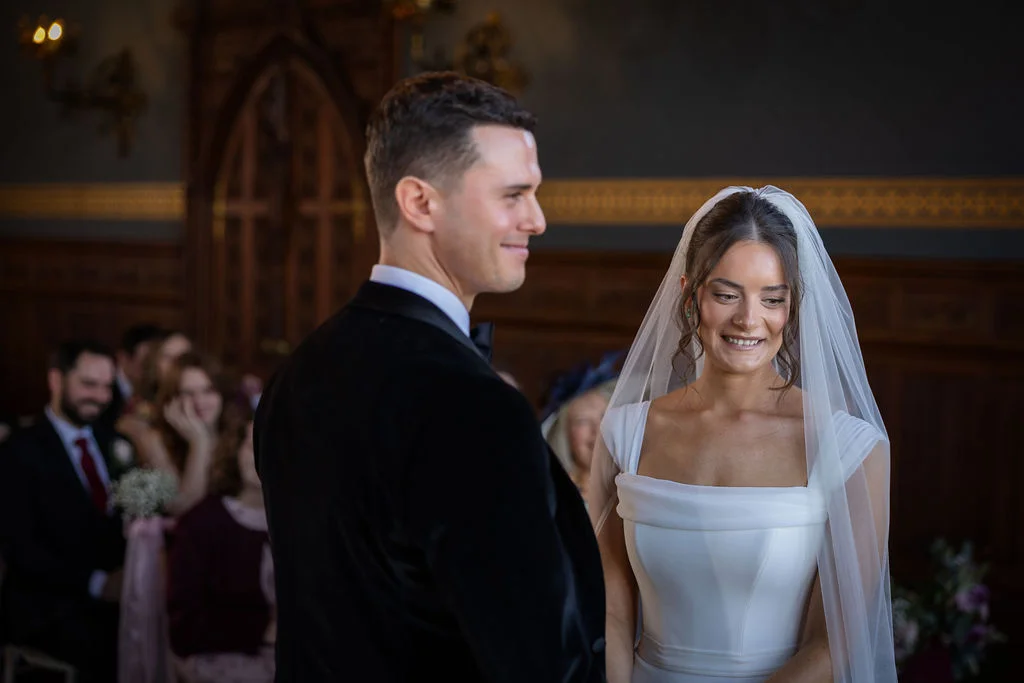 A smiling bride in a square-neck satin gown and veil stands beside her groom during an indoor ceremony inside a wood-panelled room.