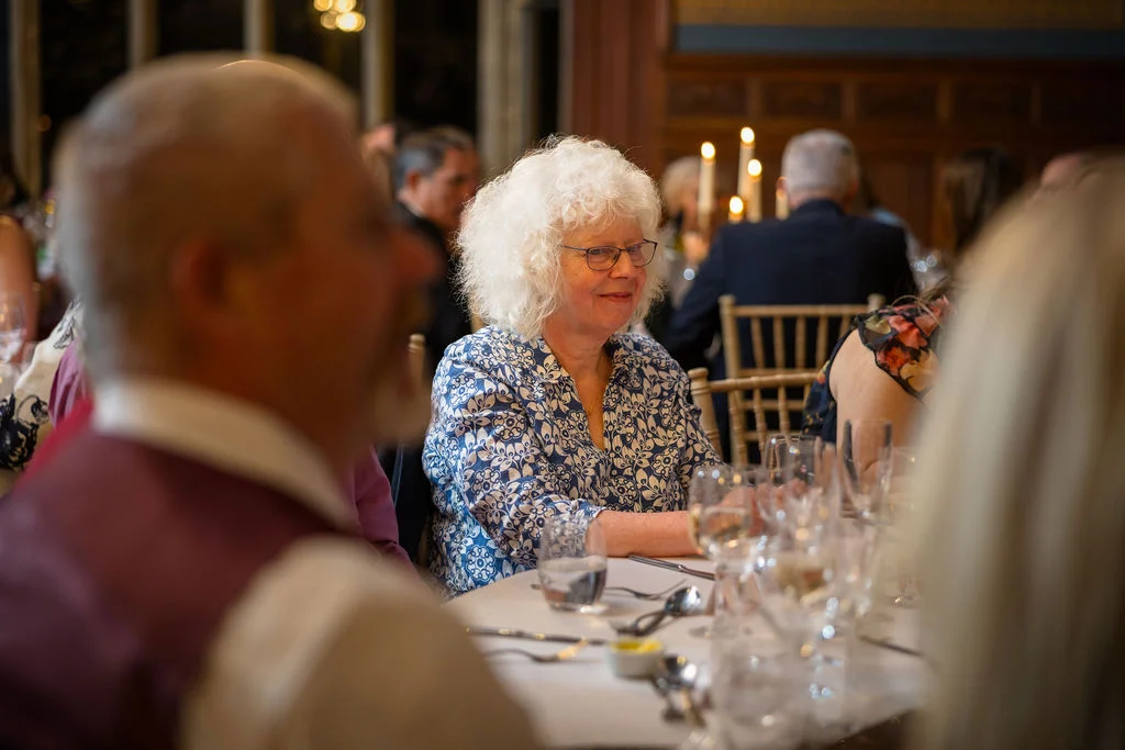 An older woman with curly white hair wearing a patterned blue and white blouse is seated at a wedding breakfast table, smiling softly amidst guests.