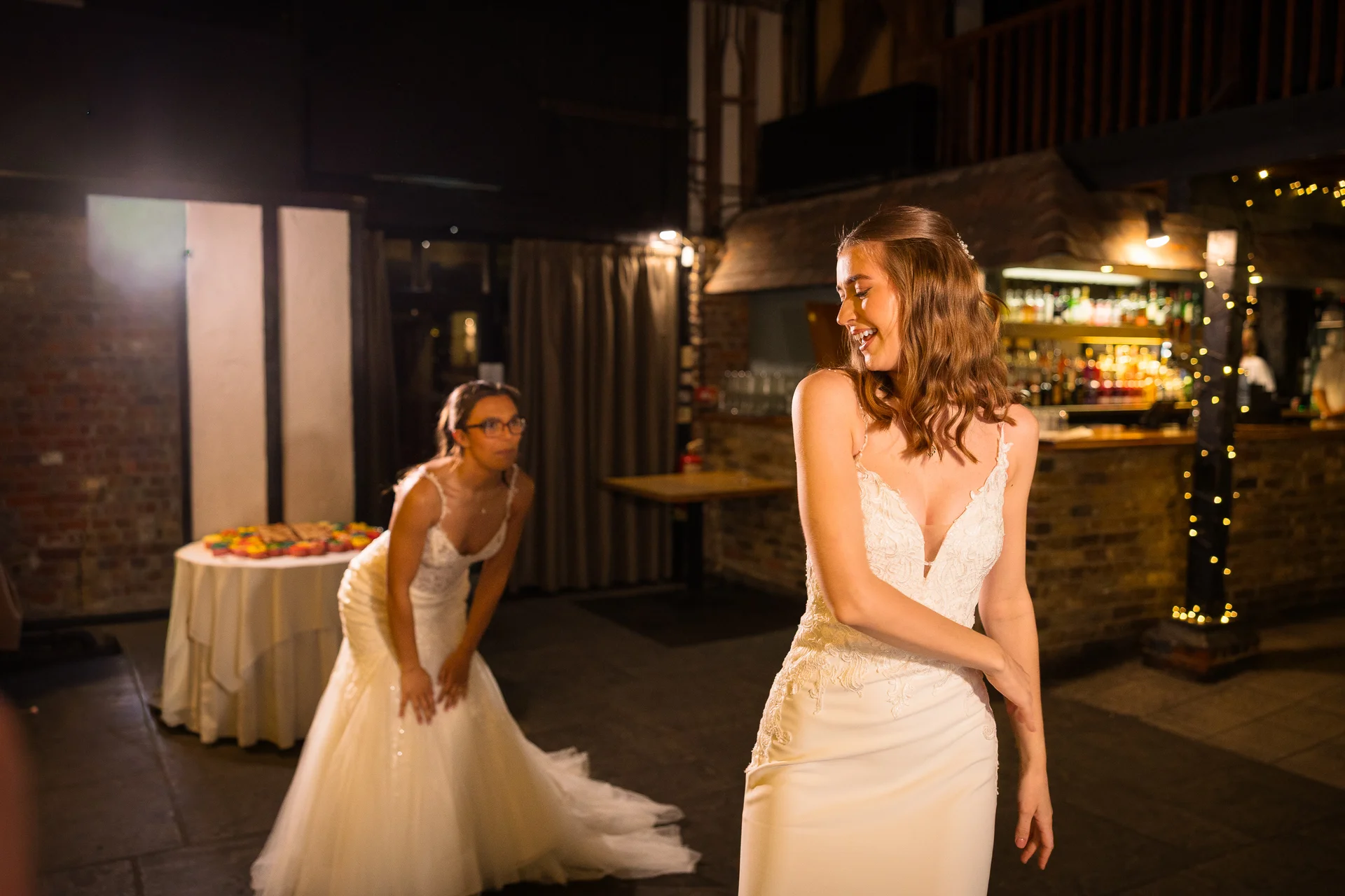 Two smiling brides share a joyful, candid moment inside a rustic wedding venue with an illuminated bar in the background.