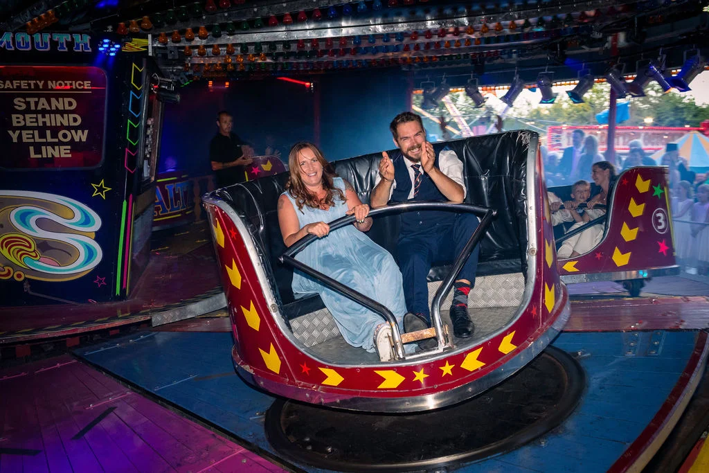 A joyful couple laughing while riding a spinning fairground attraction at an evening wedding reception celebration.