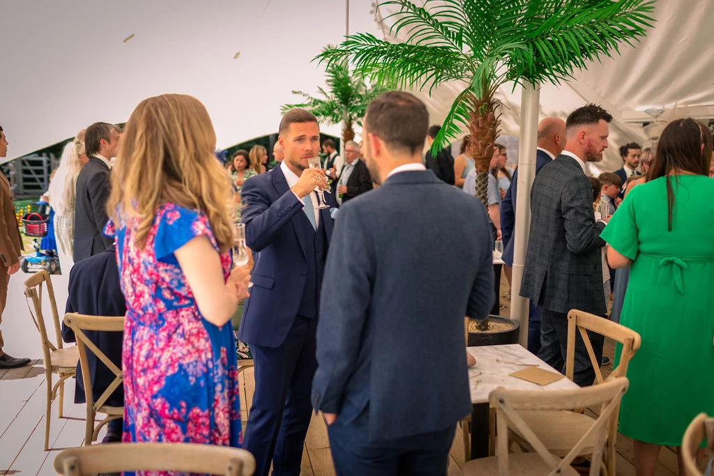 Guests enjoy drinks and conversation during a lively reception inside a bright marquee tent decorated with tropical palm trees.