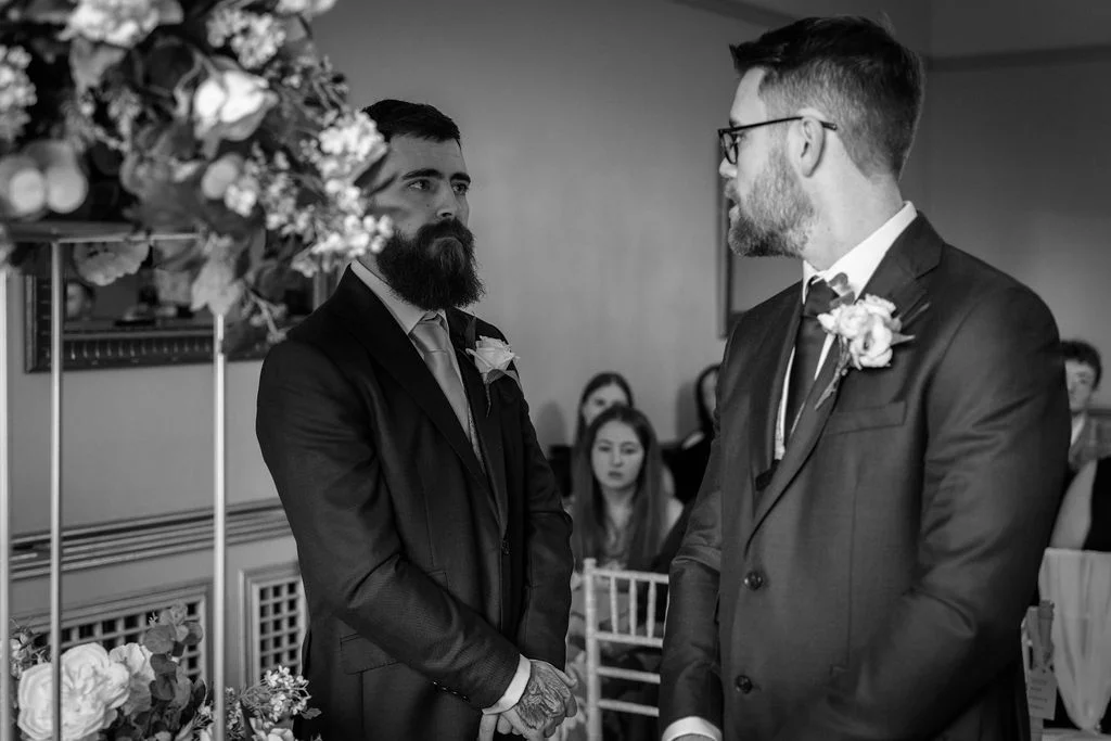 Two grooms stand facing each other during their wedding ceremony, wearing matching suits with boutonniere floral pins in a black and white capture.