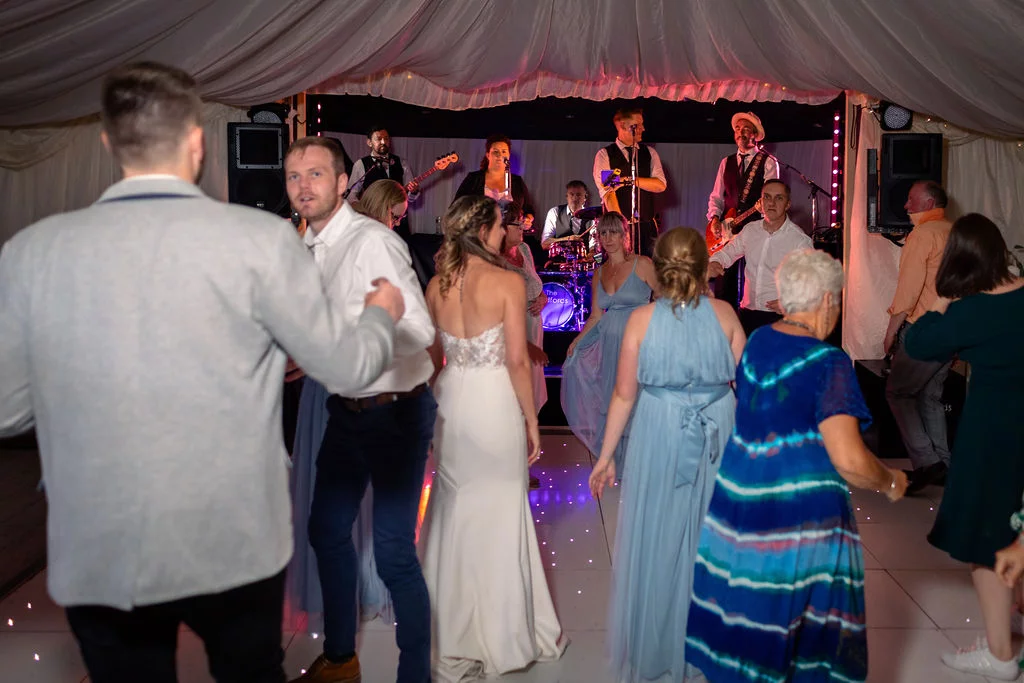 Wedding guests and the bride and groom dance energetically on a lit dance floor inside a draped marquee with a live band performing on stage.