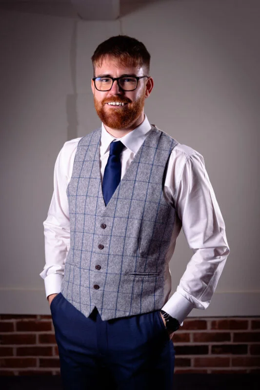 A smiling groom poses confidently in a grey checked tweed waistcoat, blue trousers, and a blue tie against a brick wall backdrop.