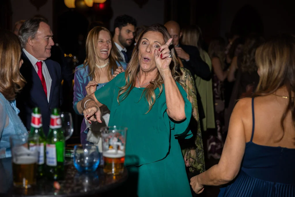 A joyful wedding guest wearing a green dress dances energetically near a crowded bar during the evening reception.
