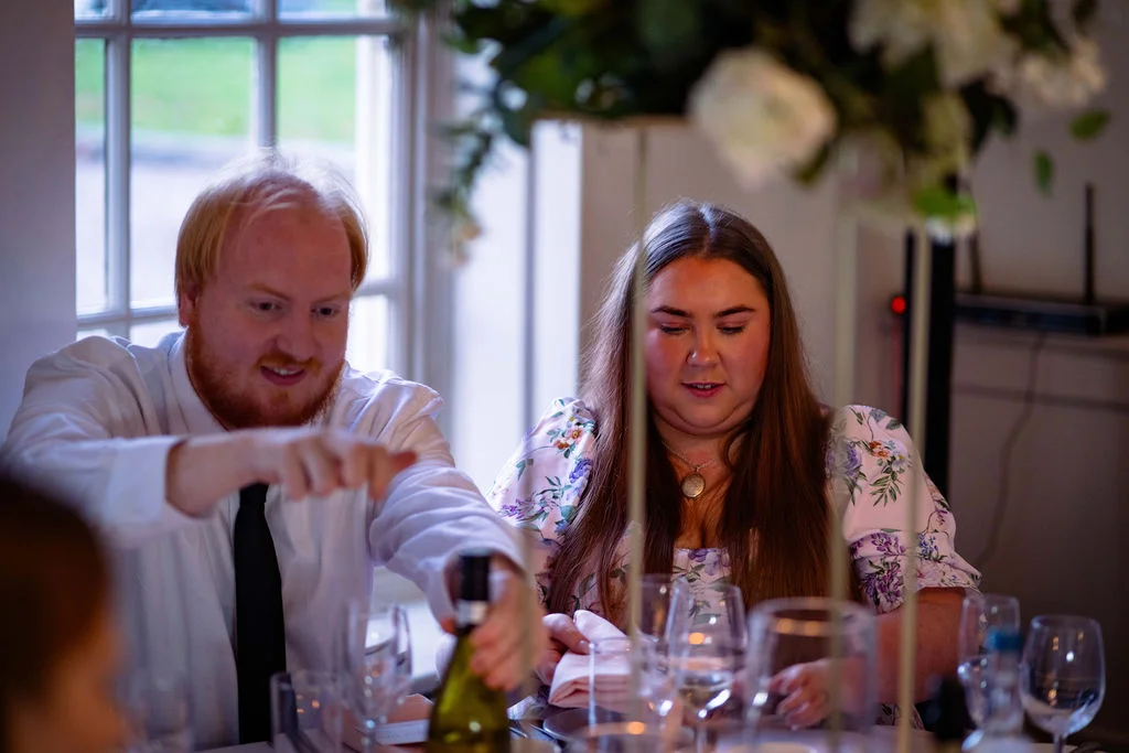 A wedding guest points towards a wine bottle at a beautifully set reception table next to another guest seated near a window.