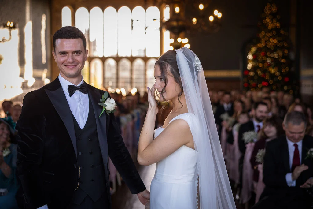 A joyful groom smiles proudly while his tearful bride gently wipes away an emotional tear during their winter wedding ceremony inside a grand hall.