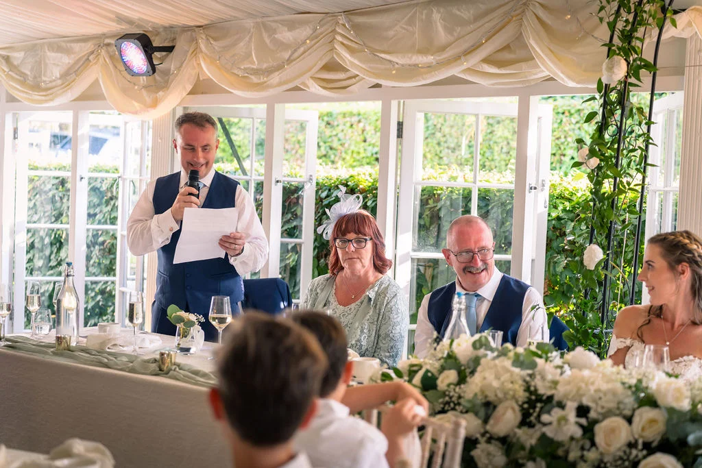A smiling guest delivers a speech holding notes at a wedding reception inside a marquee draped with white fabric and decorated with white floral centrepieces.