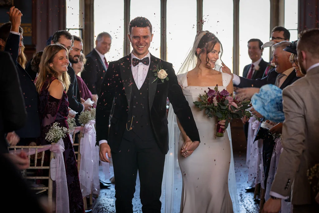 A joyful bride and groom walk hand in hand down the aisle amidst a shower of confetti, surrounded by cheering guests.