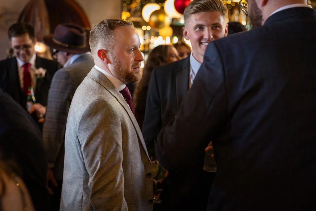 A candid capture of wedding guests in smart suits mingling and chatting during the drinks reception.