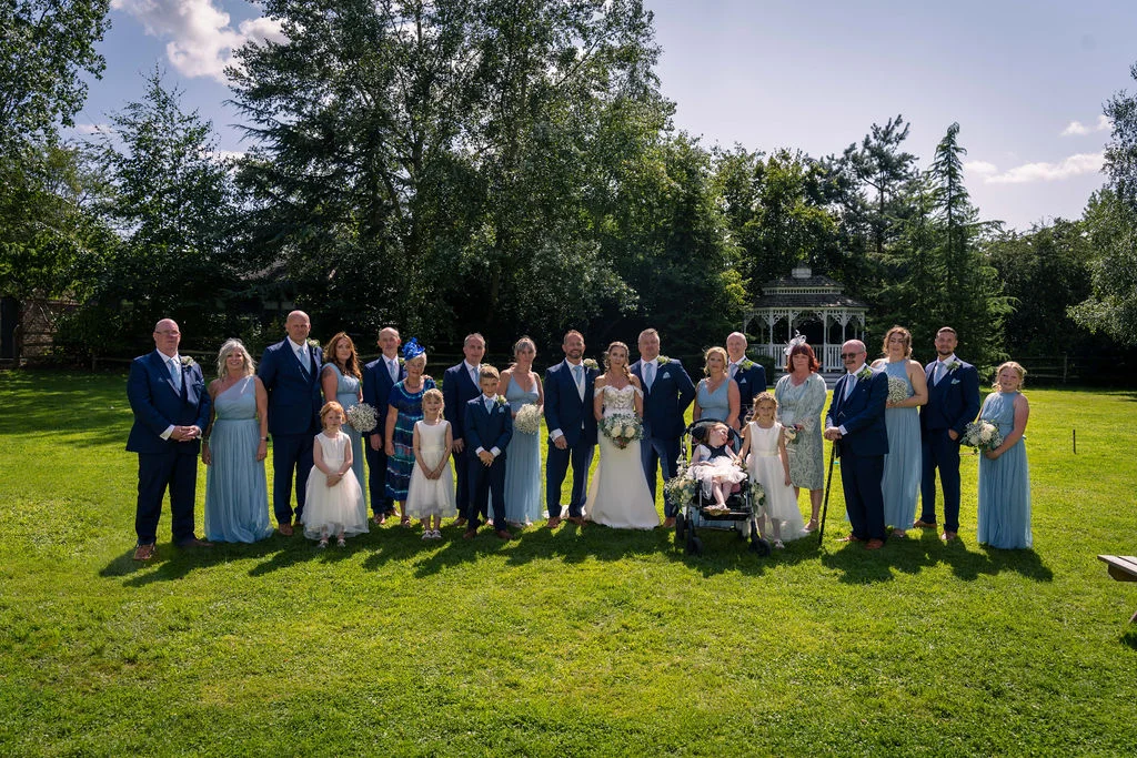 A large wedding party and family group pose for a formal portrait outdoors on a sunny lawn in front of a white gazebo.