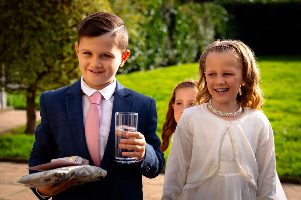 A young page boy in a navy suit and pink tie holds a glass of water and wrapped gifts, walking alongside a smiling flower girl wearing a white dress and cardigan outdoors.