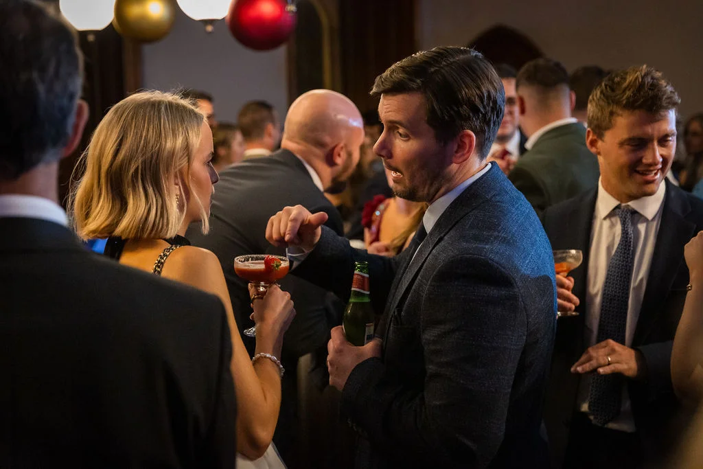 Wedding guests mingle and chat animatedly during a lively evening drinks reception while holding cocktails and bottled drinks.