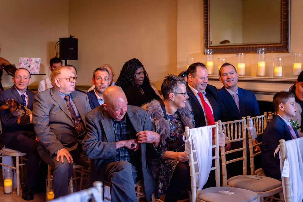 Wedding guests are seated in a candlelit room during a ceremony, smiling and watching attentively.