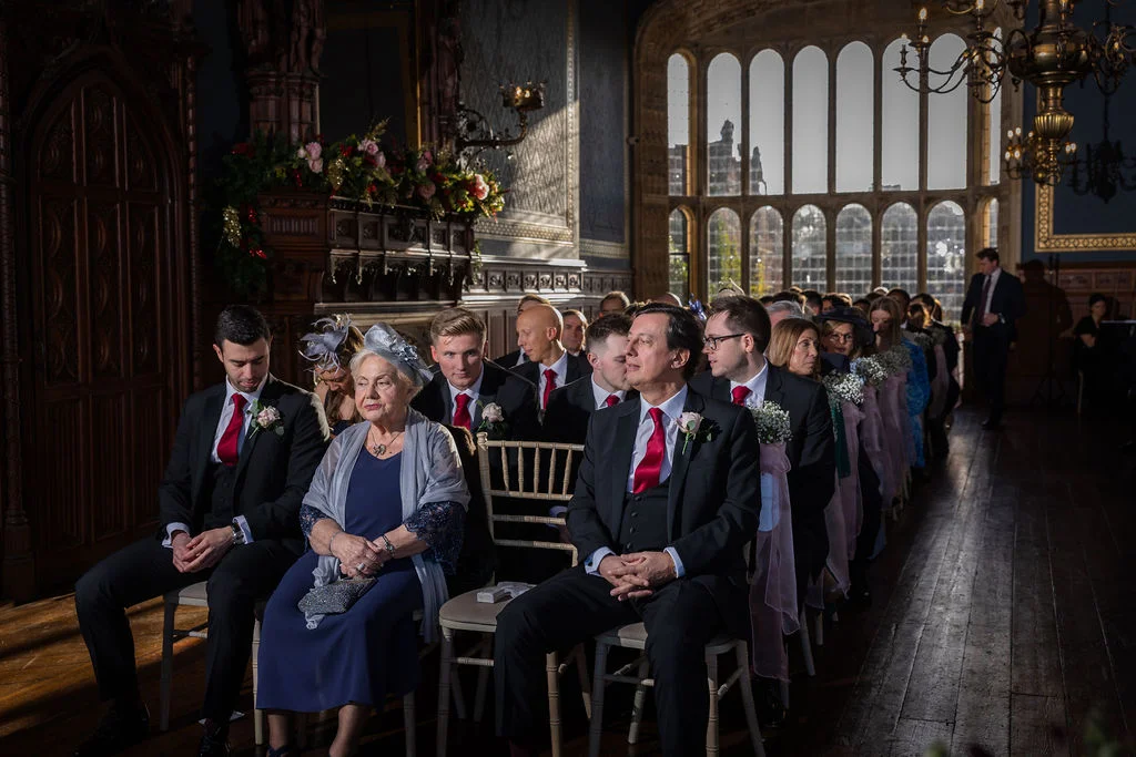 Wedding guests seated in rows along the aisle inside a grand historic hall featuring tall arched windows and dramatic lighting.