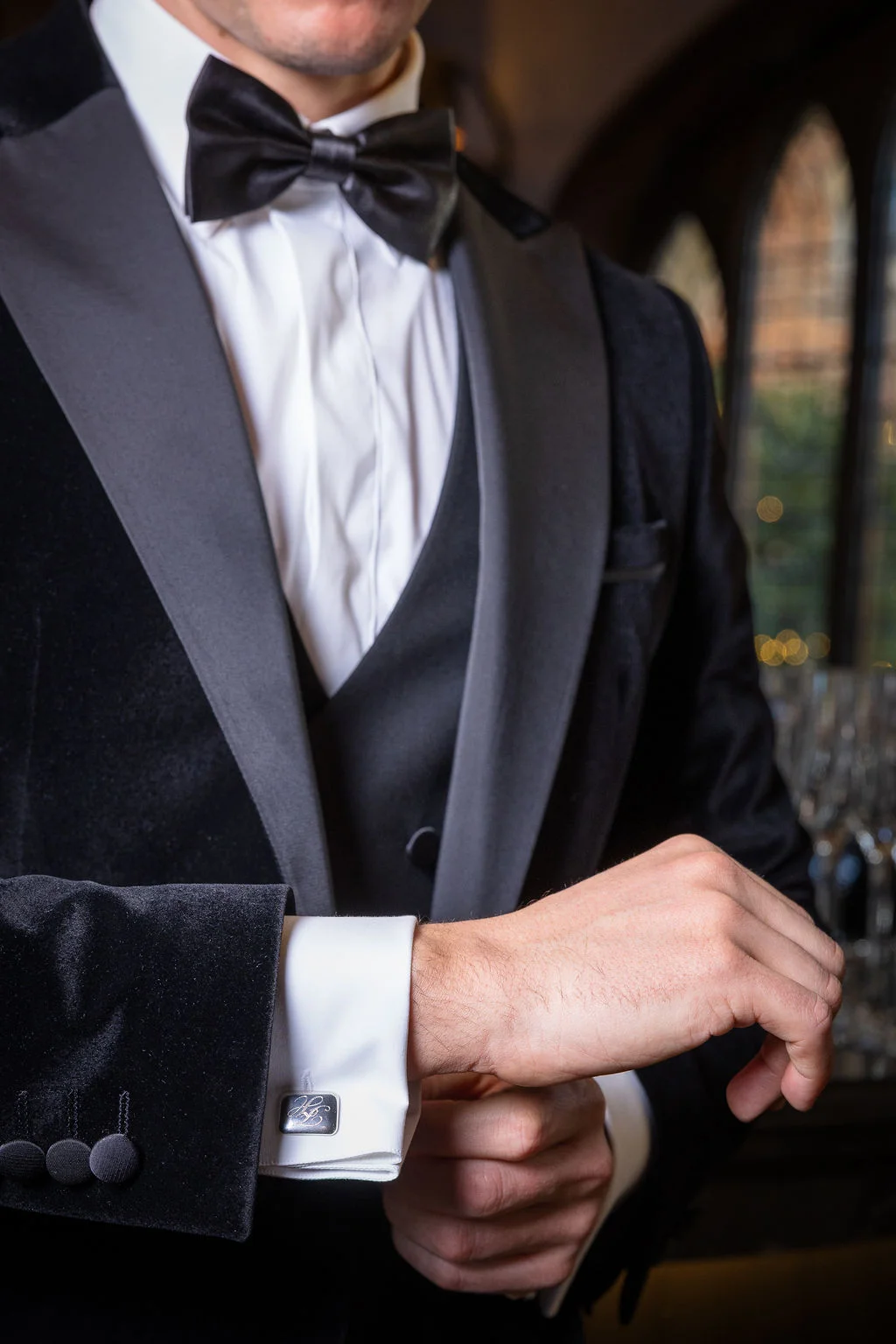 A groom dressed in a sophisticated black velvet tuxedo and bow tie adjusts his engraved cufflinks while getting ready for the wedding.