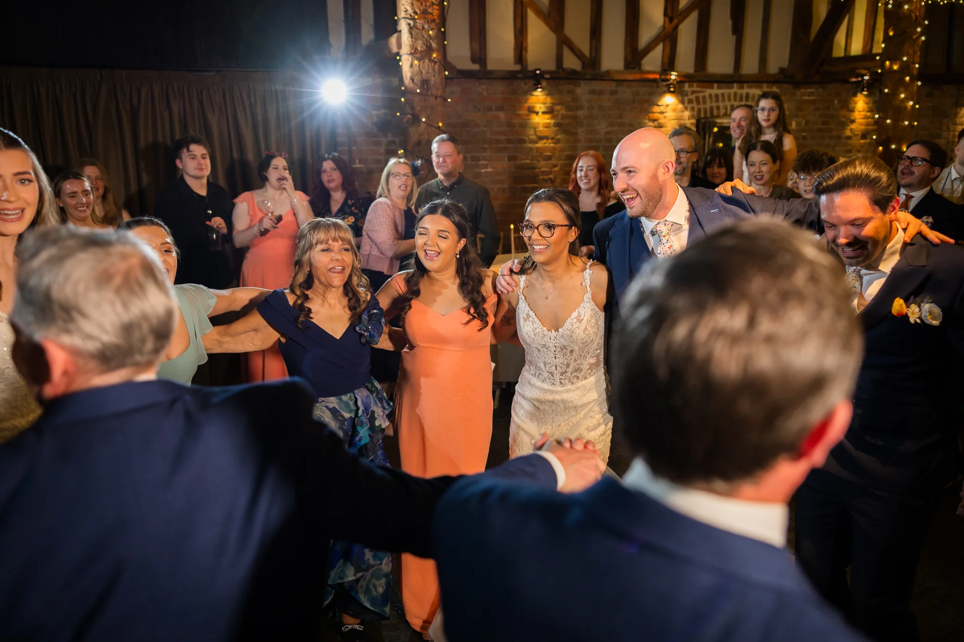 A joyful group of wedding guests and the newlyweds form a lively dance circle inside a rustic barn with exposed brick and warm string lighting.