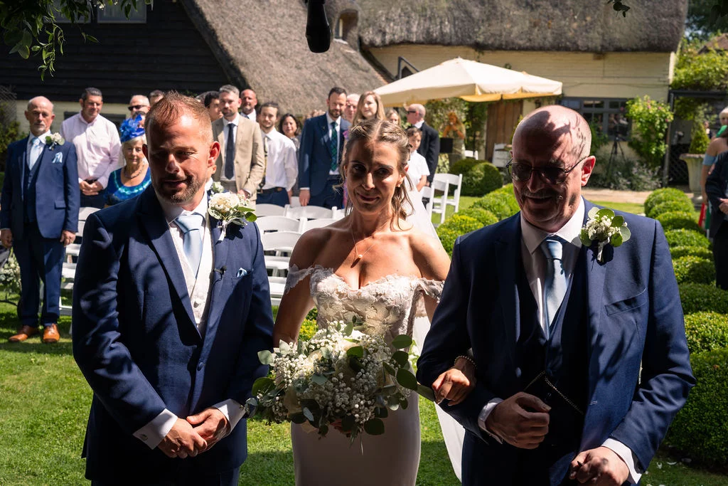 A smiling bride walks down an outdoor garden aisle linked arm in arm with her father towards the waiting groom during a sunny summer ceremony.