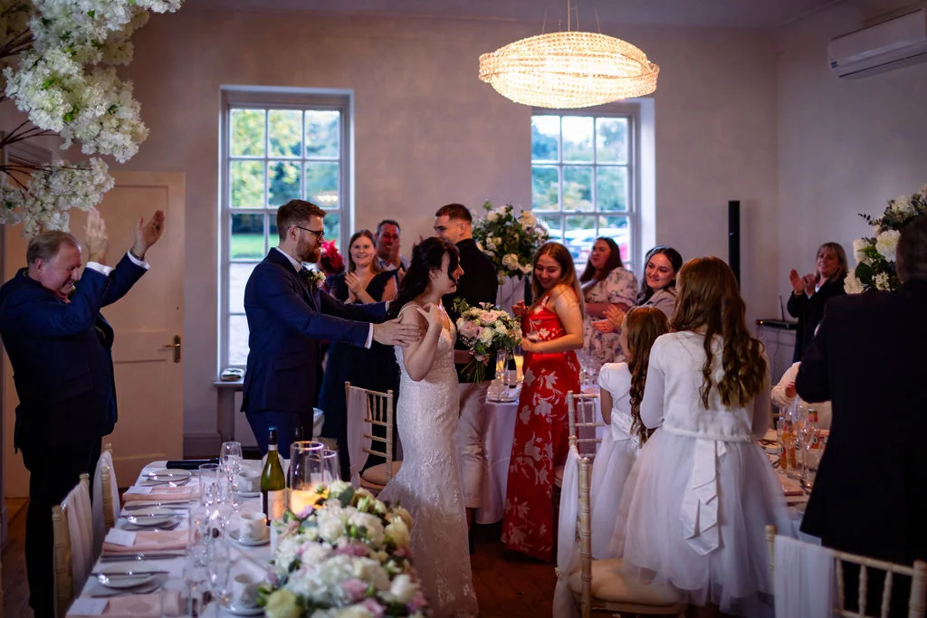 The newlywed couple enter their wedding reception to applause and cheering from guests seated at elegantly decorated tables.