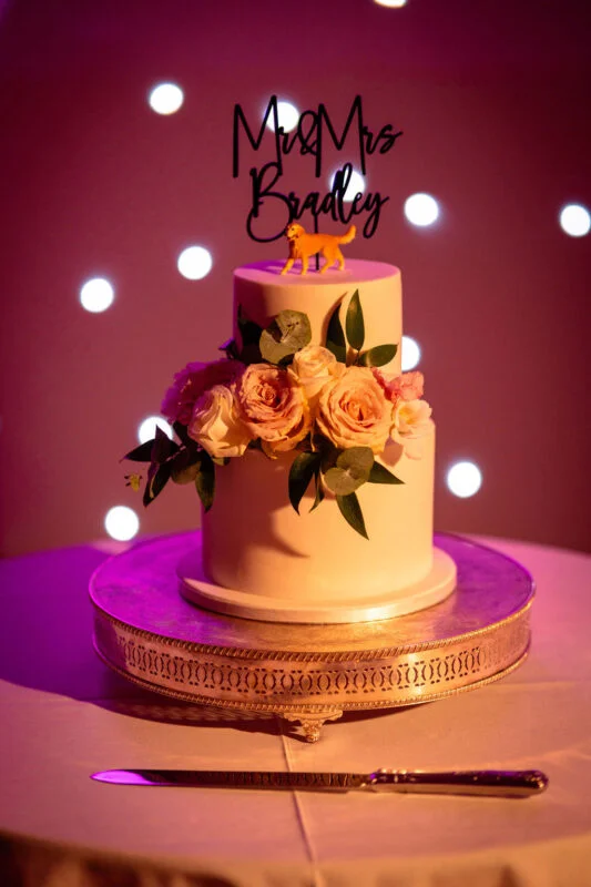 A two-tier wedding cake adorned with fresh roses and eucalyptus sits on an ornate silver stand against a backdrop of soft fairy lights.