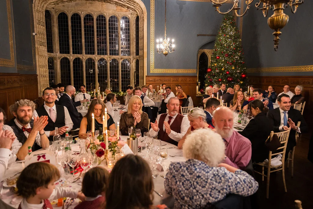 Wedding guests enjoy speeches and dinner in a grand panelled banquet hall decorated with a festive Christmas tree and candlelight.