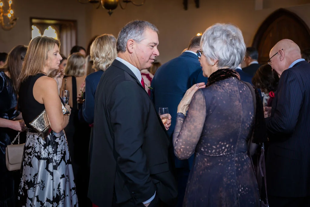 Wedding guests dressed in formal evening attire mingle and converse during a drinks reception inside a venue with warm interior lighting.