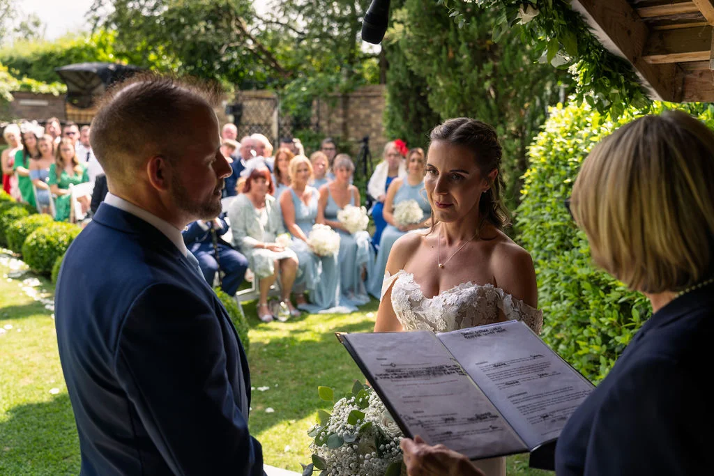 A bride and groom stand outdoors in a lush garden during a celebrant-led ceremony, with guests seated in the background as they participate in the wedding proceedings.