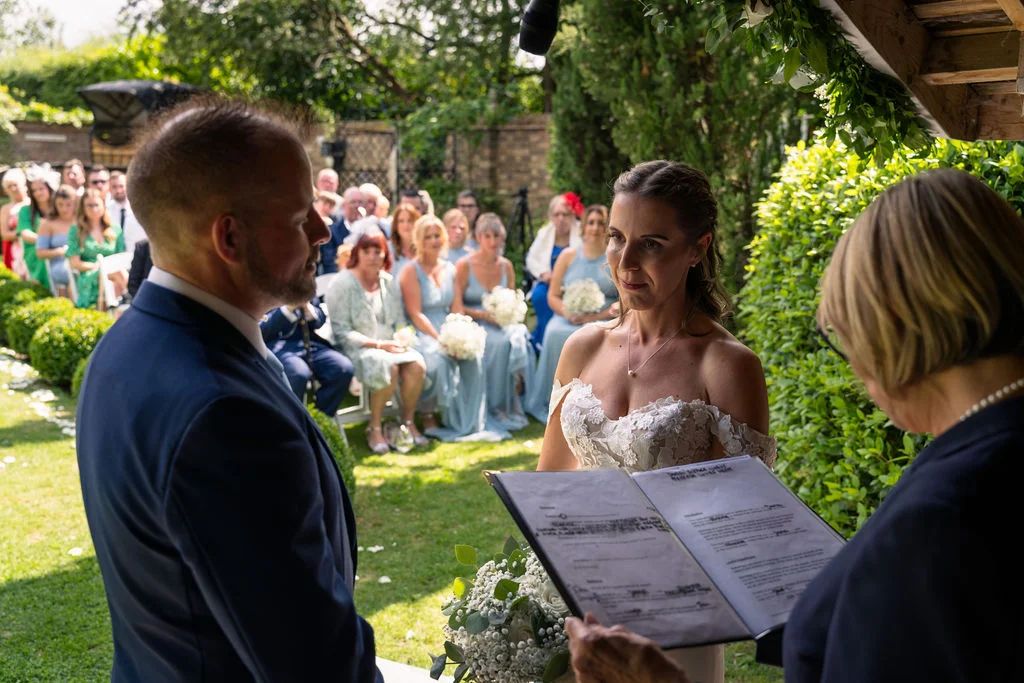A bride and groom stand during an outdoor garden wedding ceremony while a celebrant holds a service book in front of seated guests.