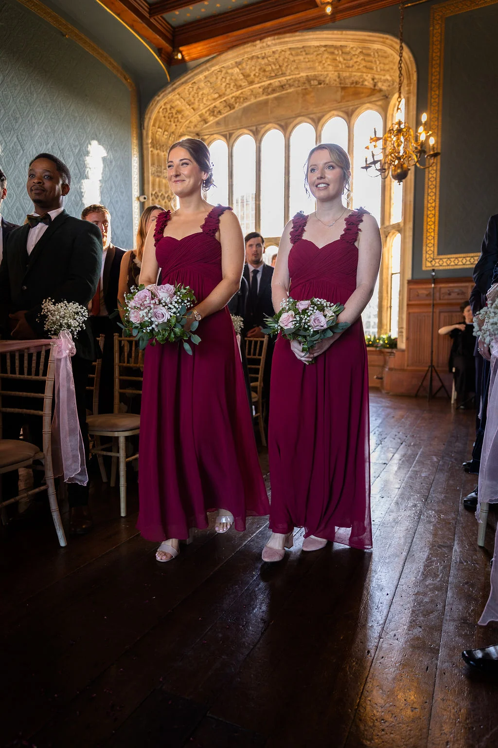 Two smiling bridesmaids in matching floor-length burgundy dresses walk down the wooden aisle holding posy bouquets during a stately home wedding ceremony.