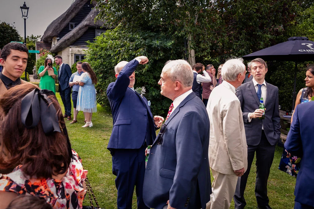 Wedding guests mingle and chat on a sunny lawn during an outdoor drinks reception near a traditional thatched building.