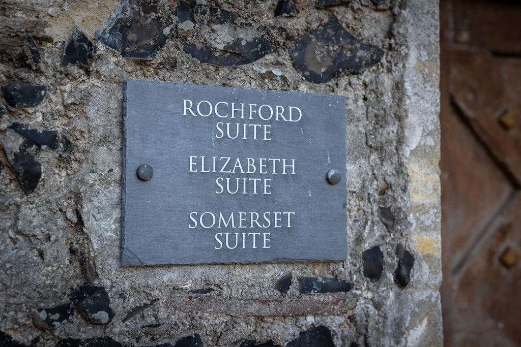 A close up view of a dark slate venue sign mounted on a rustic stone wall, pointing guests towards various suites.