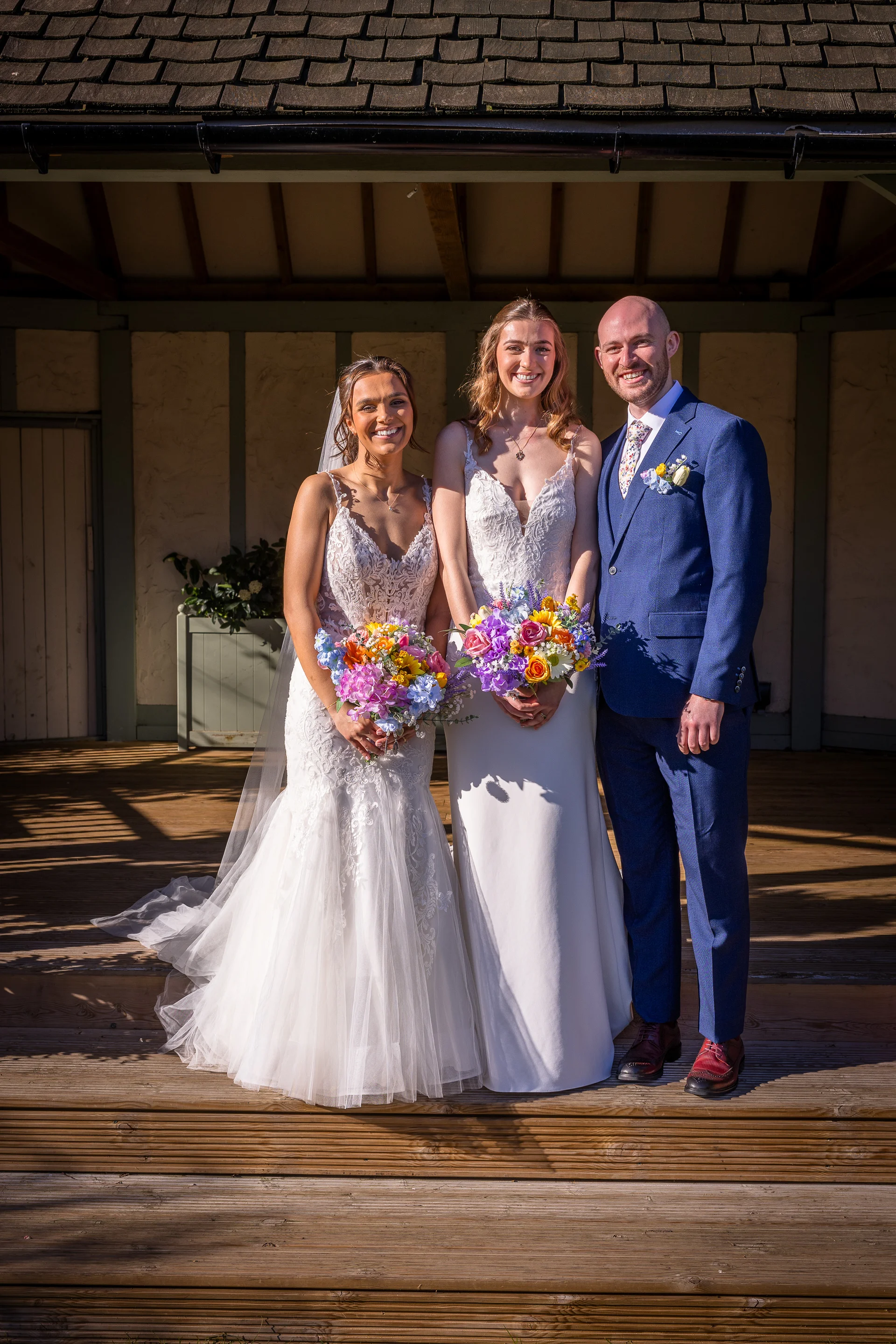 A smiling wedding party featuring two brides in lace-accented white wedding gowns and a groom in a blue suit stand together on wooden decking outdoors, holding vibrant colourful bouquets.