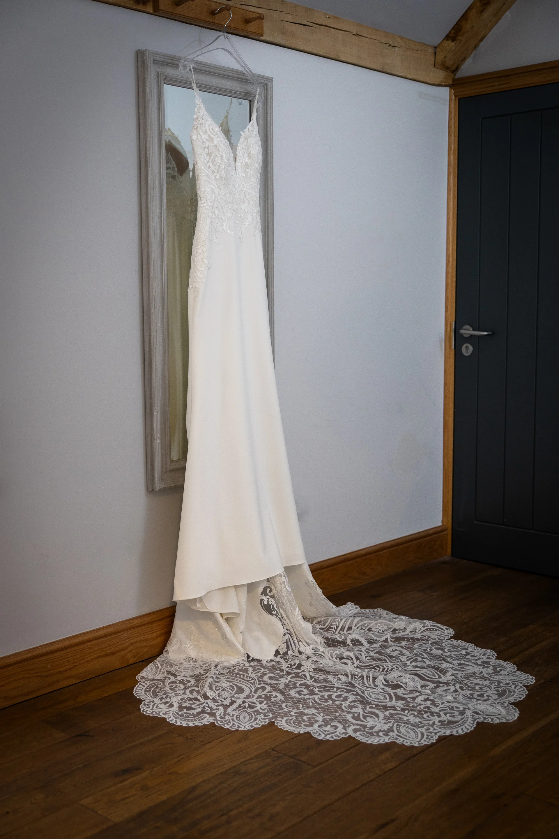 An elegant white lace wedding dress hangs delicately on a mirror inside a bright room with wooden flooring during morning bridal preparations.