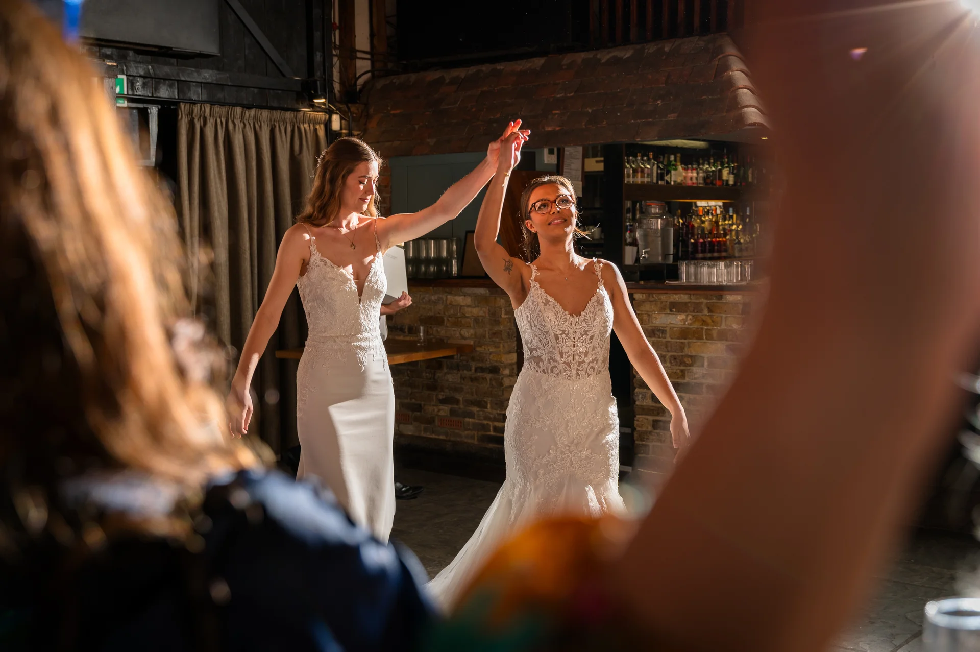 Two brides share their first dance together inside a rustic pub venue framed by guests enjoying the celebration.