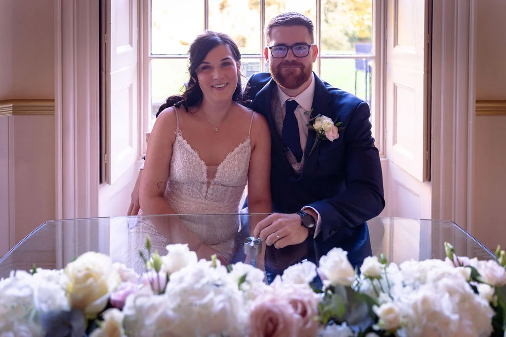A newlywed couple smiling together behind a glass table adorned with white and pink floral arrangements near a bright window.