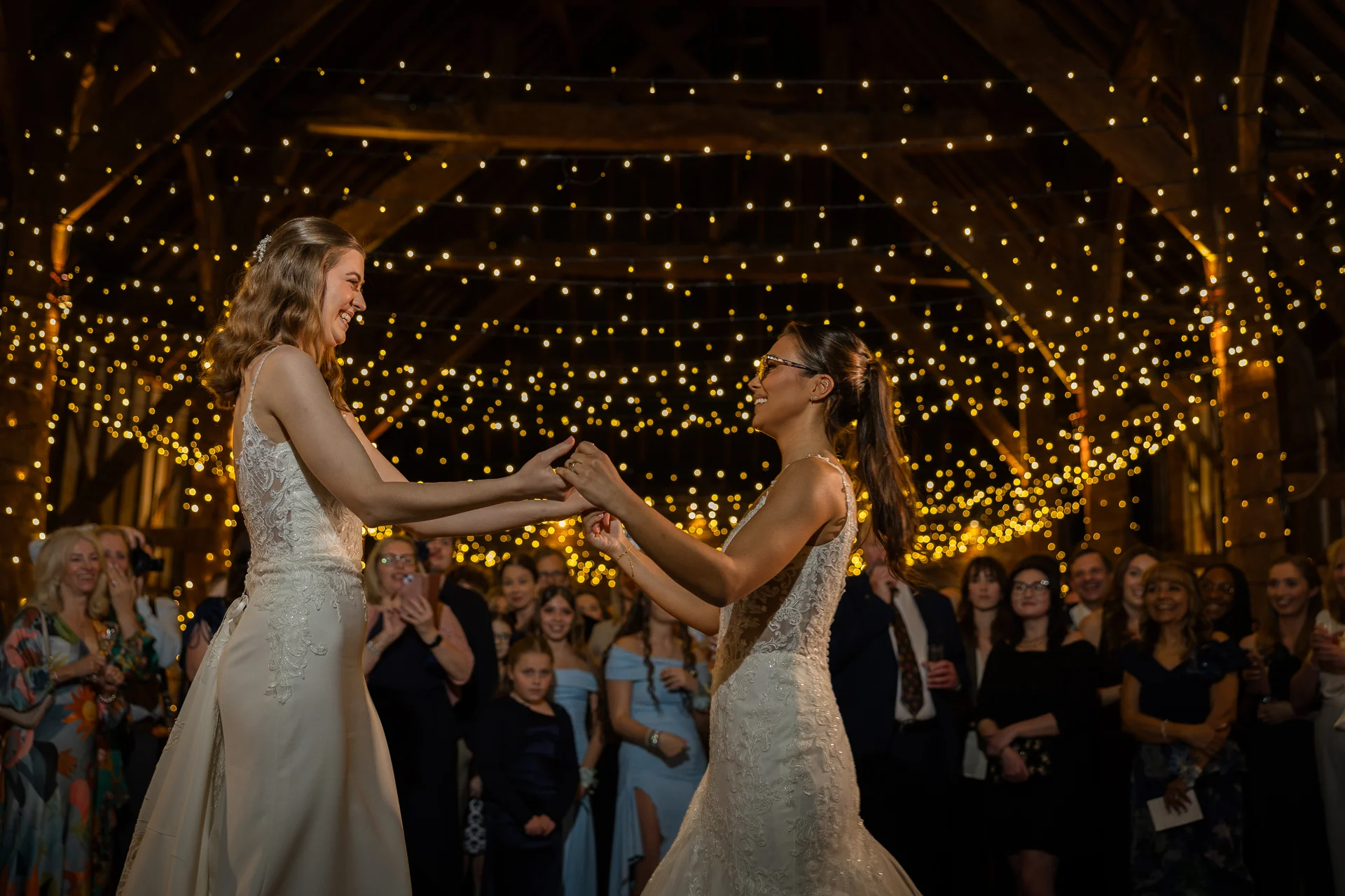 Two brides joyfully hold hands during their first dance under a breathtaking canopy of fairy lights inside a rustic wooden barn.